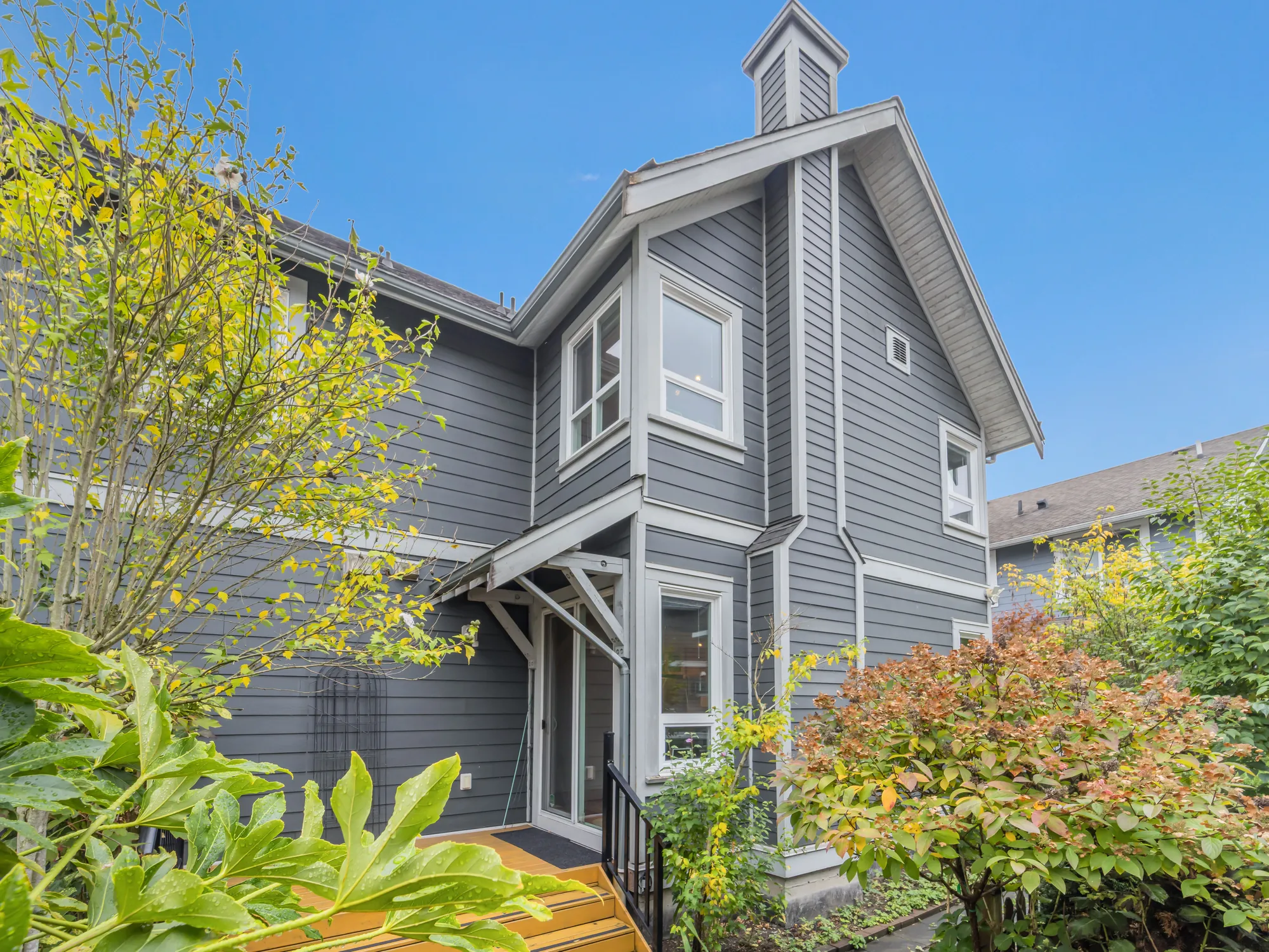The back of the house: grey siding, a chimney chase with a white cap, windows on both floors, a door onto the back deck, trees and shrubs in autumn leaf