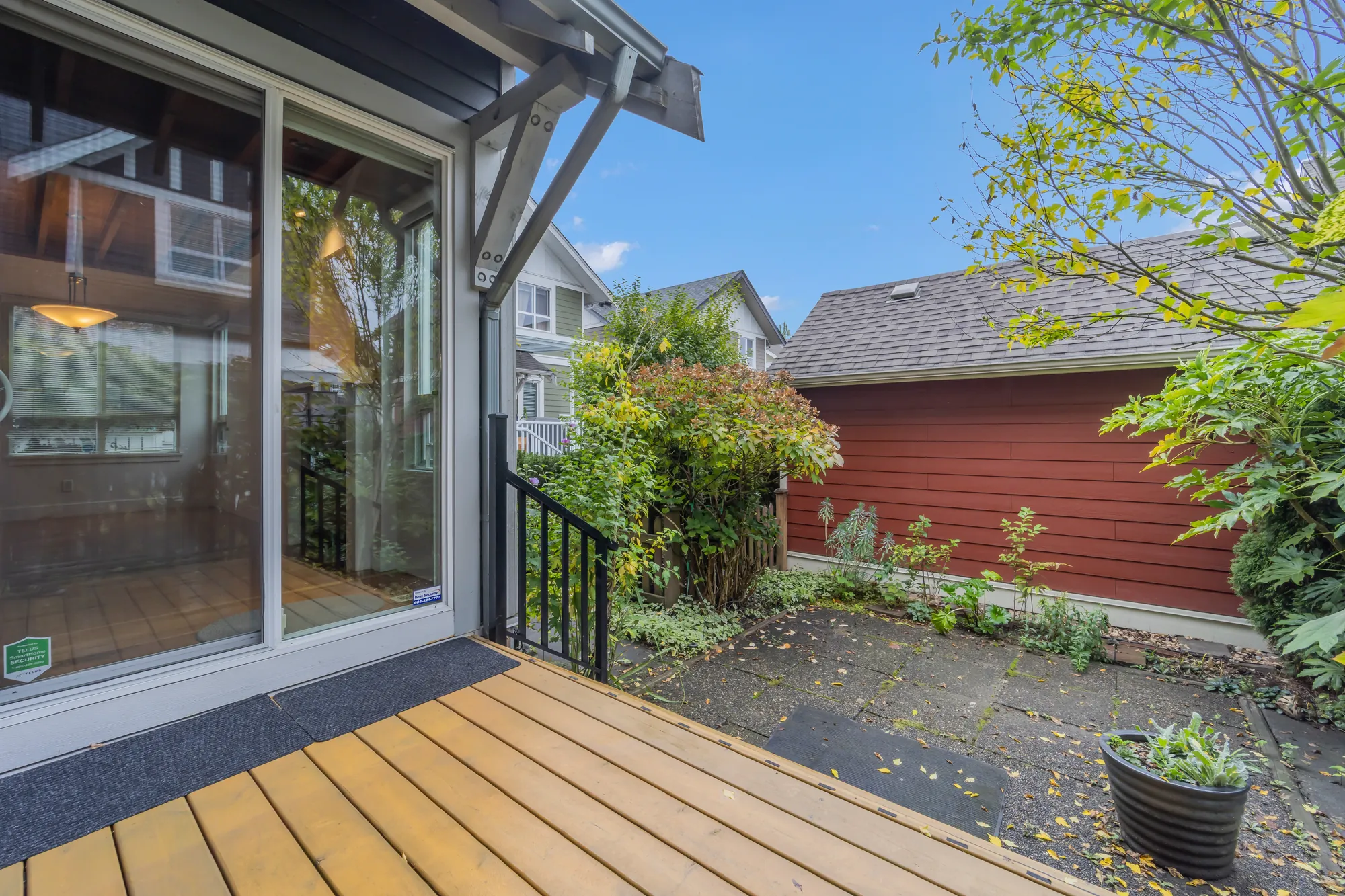 The back deck at a sliding door: wood boards and a black railing, a paved yard beyond with a planter and a bed planted along a red-sided garage