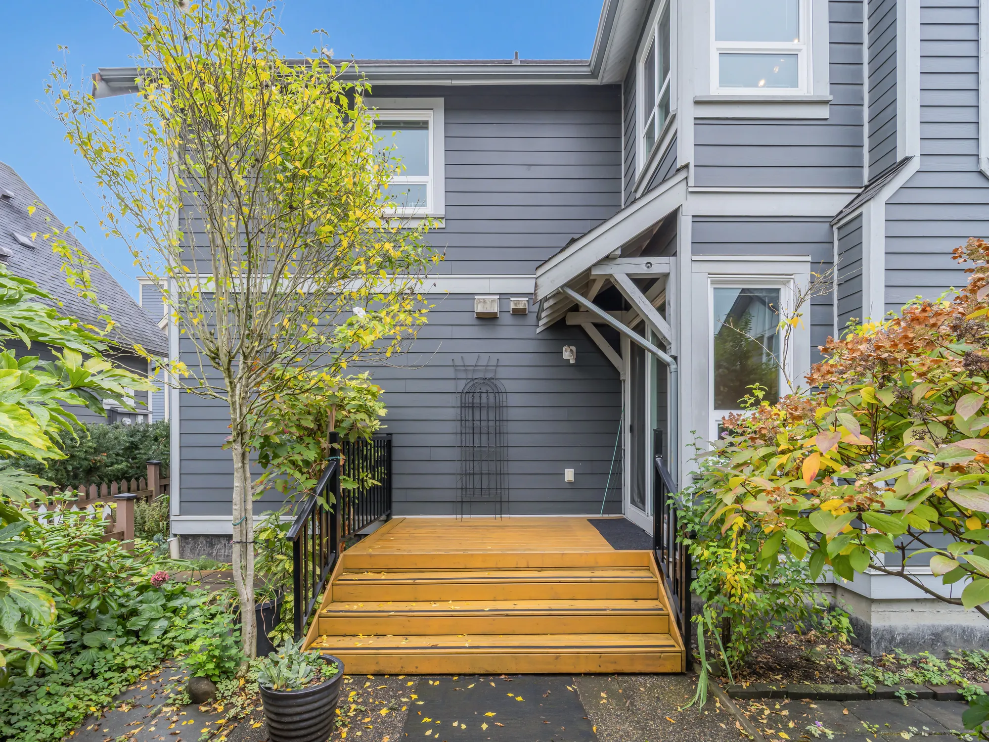 Wide wooden steps up to the back deck and a door, black railings, a young tree and shrubs in autumn colour