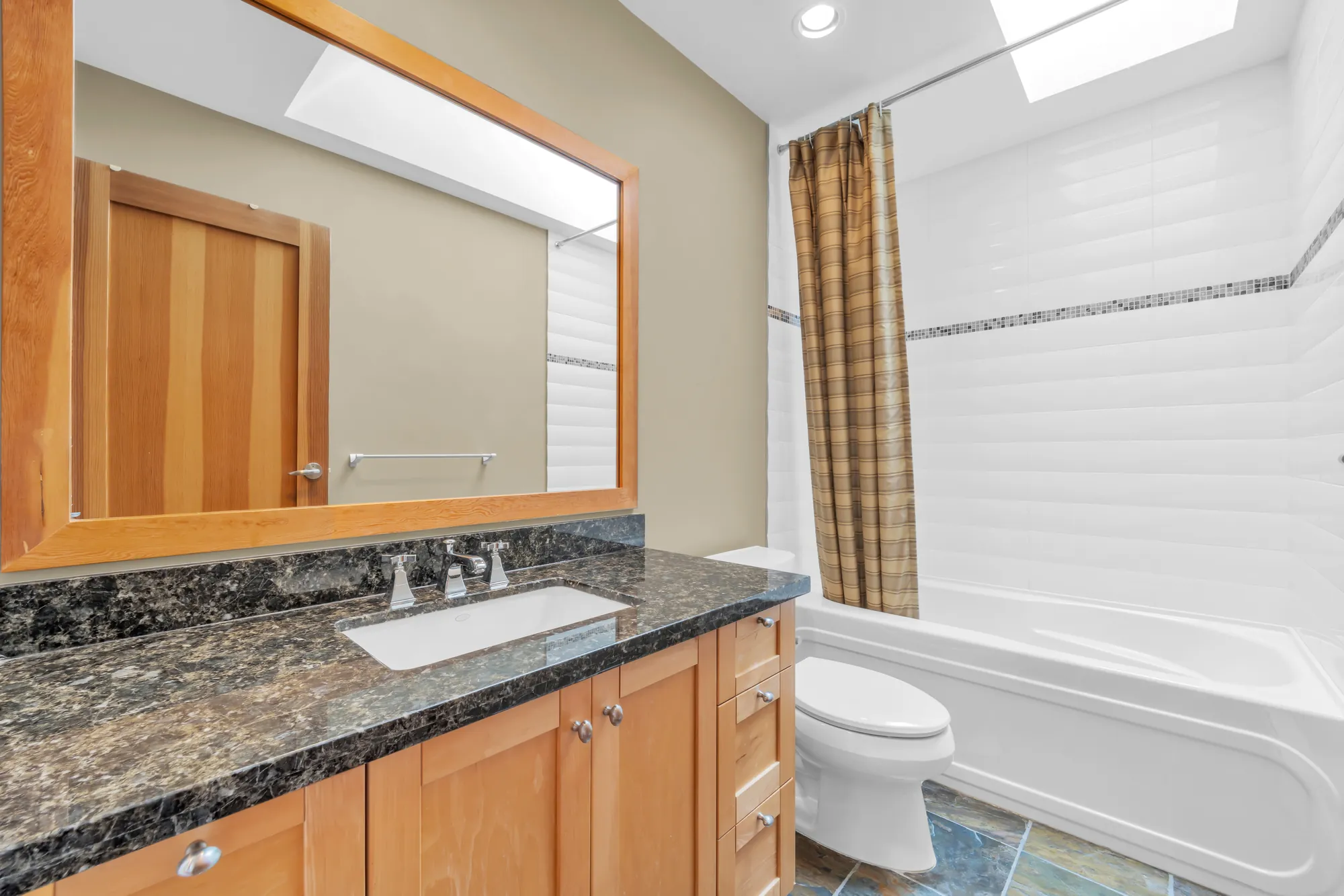 The hall bathroom: a maple vanity with a dark granite top and a wood-framed mirror, a toilet, a tub with a shower curtain, a skylight
