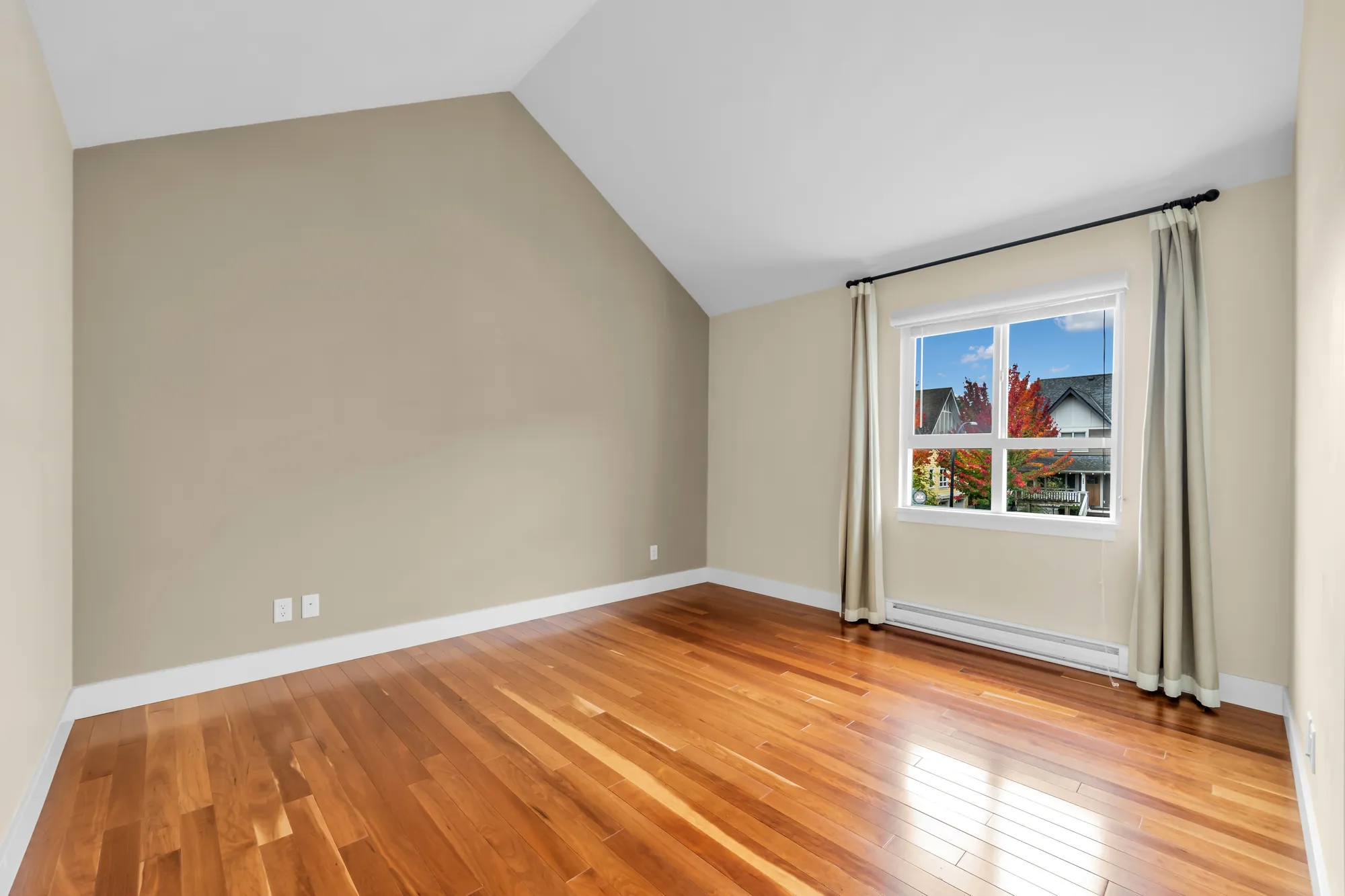 The second bedroom, empty: a vaulted ceiling, a taupe wall, a window with cream curtains, wood floors