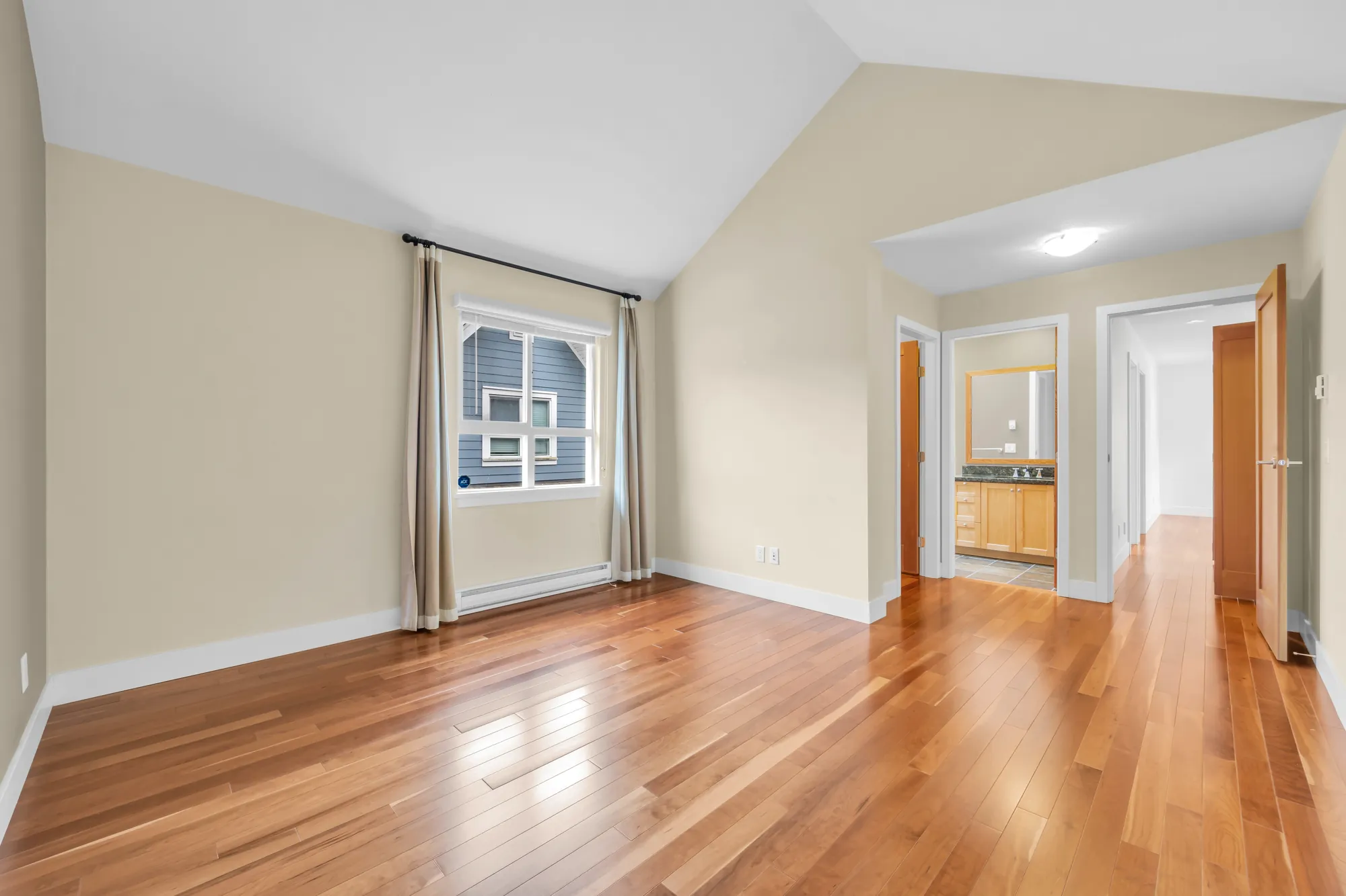 The second bedroom toward a window with cream curtains, the doorway to its bathroom and the door to the hall