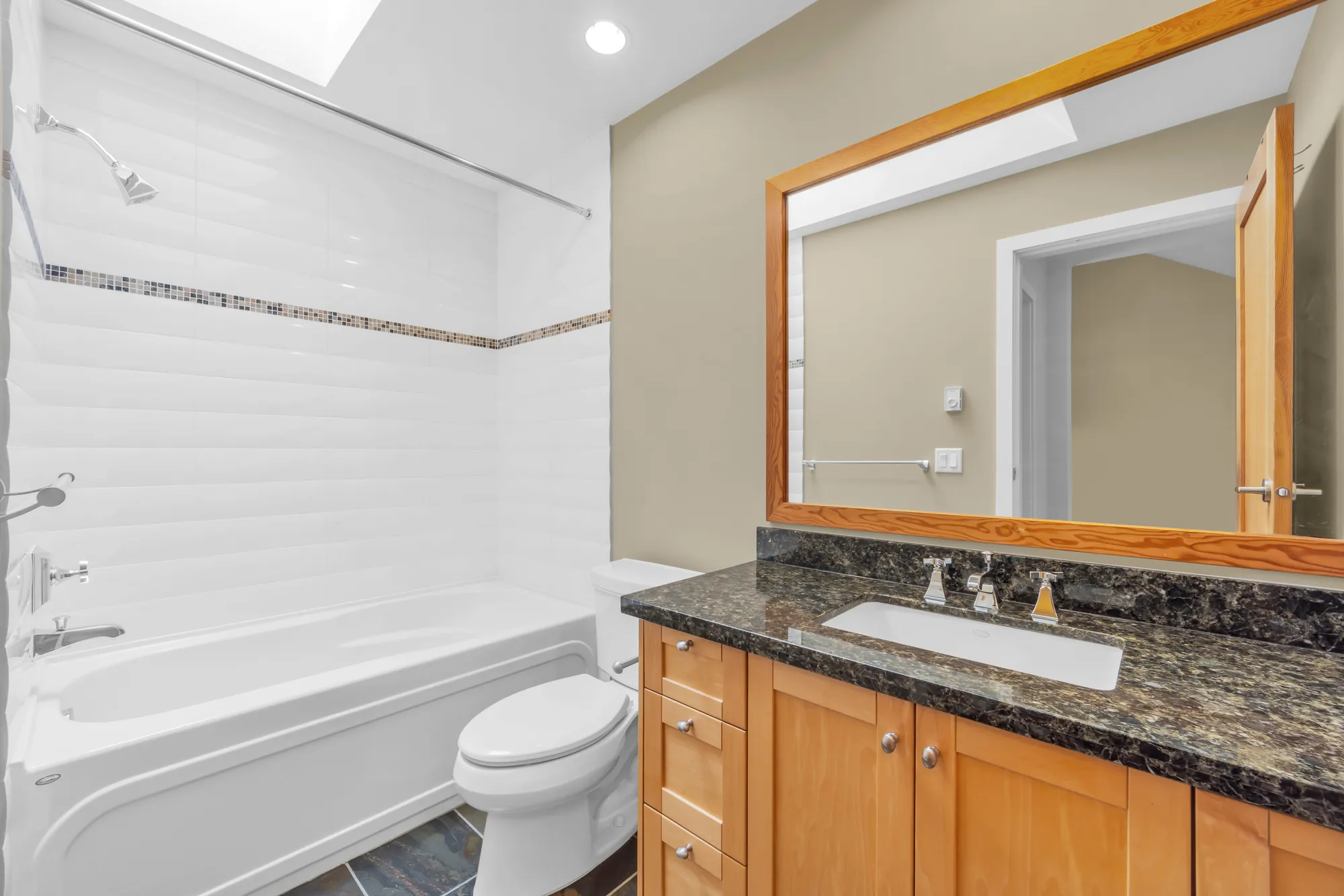 A bathroom: a tub with a shower in white tile with a mosaic band, a toilet, a maple vanity with a dark granite top, a skylight