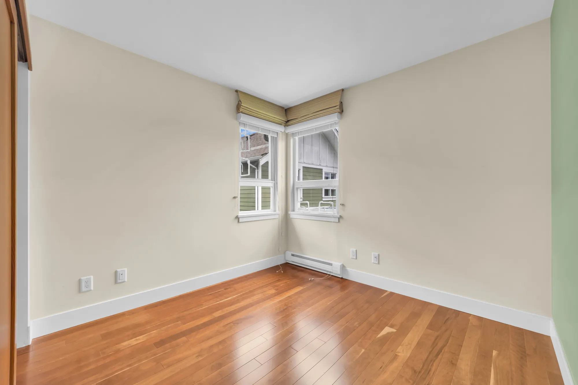 The third bedroom, empty: two windows meeting at a corner under roman shades, a sage green wall at the right, wood floors