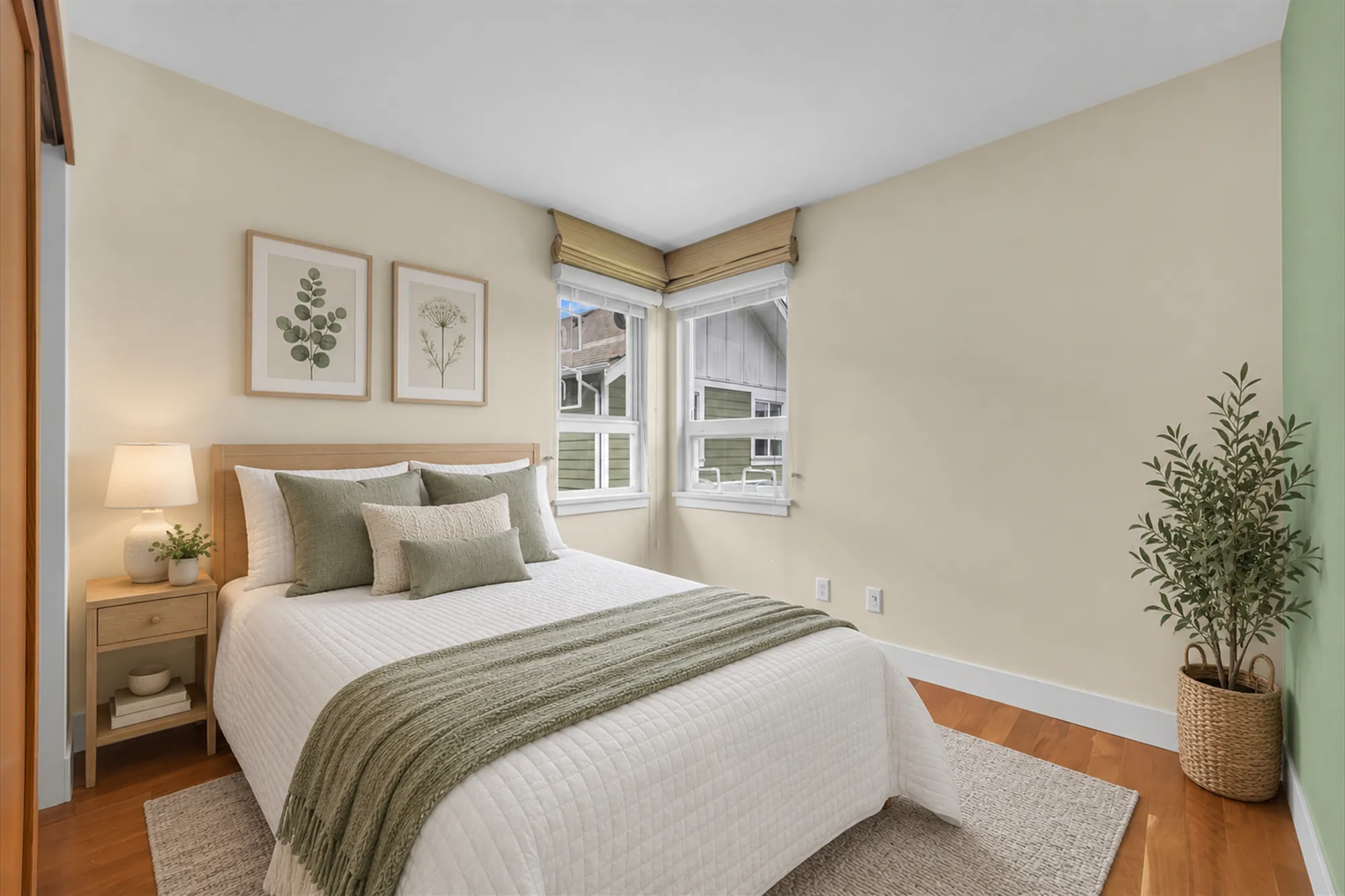 The same frame virtually staged: a bed with green and white bedding, a night table with a lamp, a rug, an olive tree in a basket, two botanical prints