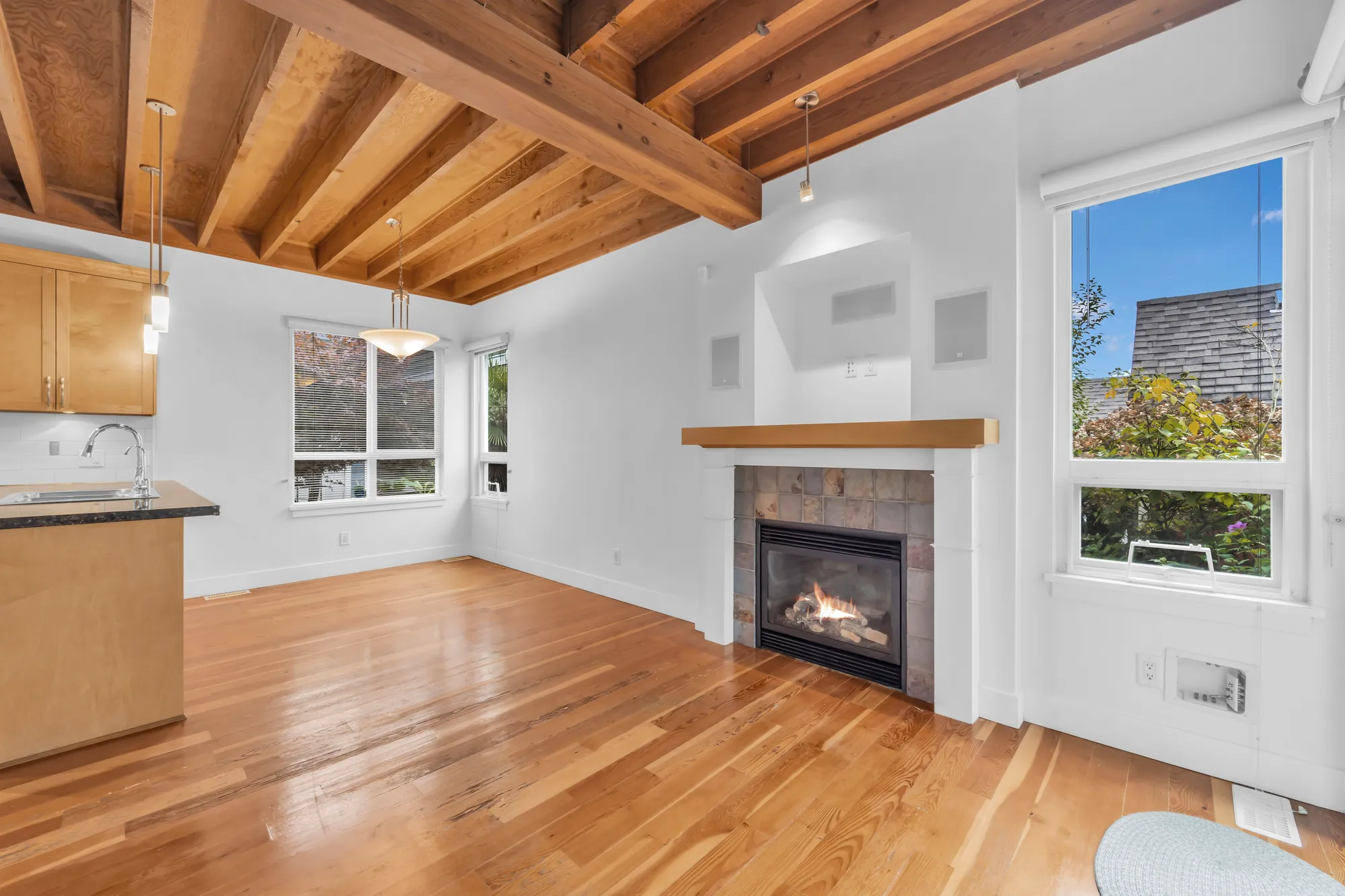 The family room and eating area, empty: exposed wood joists and a beam, a fireplace in slate tile with a wood mantel and a niche above it, a wide window, the eating area&rsquo;s window, the kitchen at the left