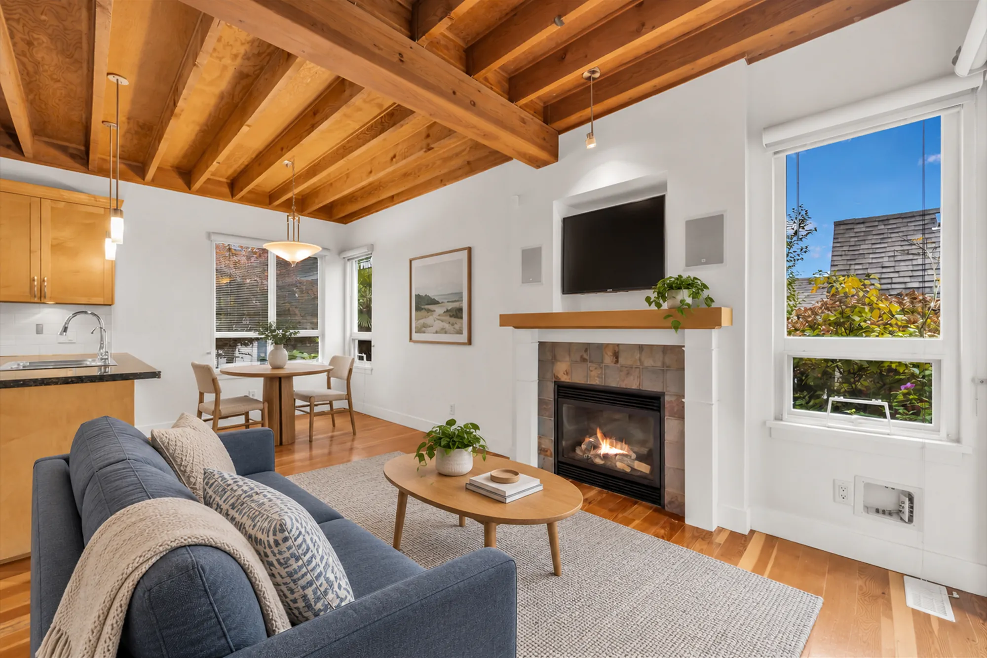 The same frame virtually staged: a blue sofa, an oval coffee table on a rug, a television in the niche over the fireplace, a print, a small round table with two chairs in the eating area, plants