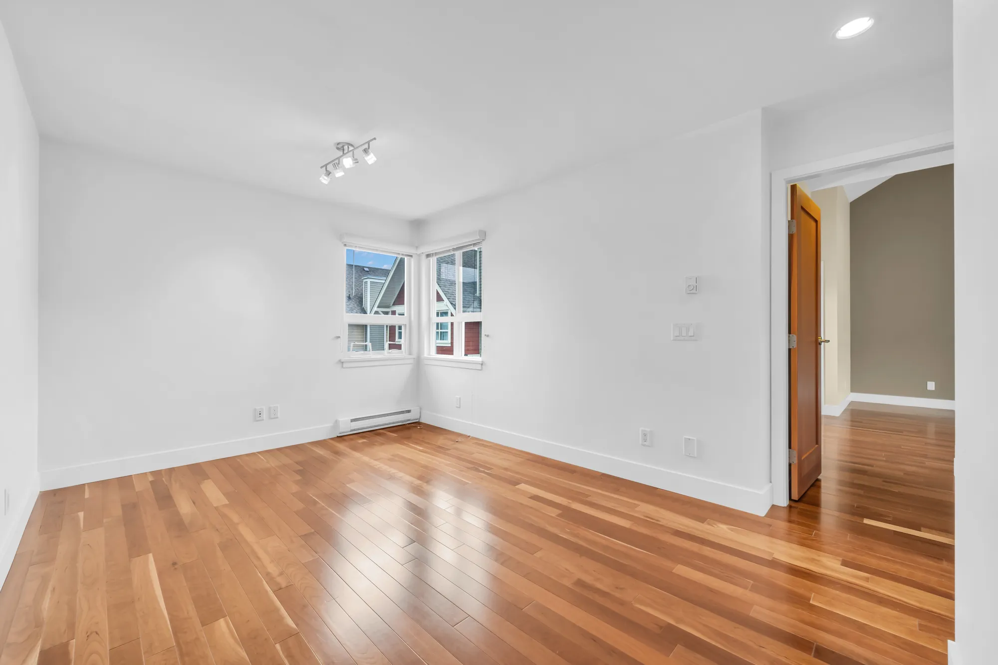 The flex room, empty: white walls, two windows meeting at a corner, track lights, wood floors, a door open at the right