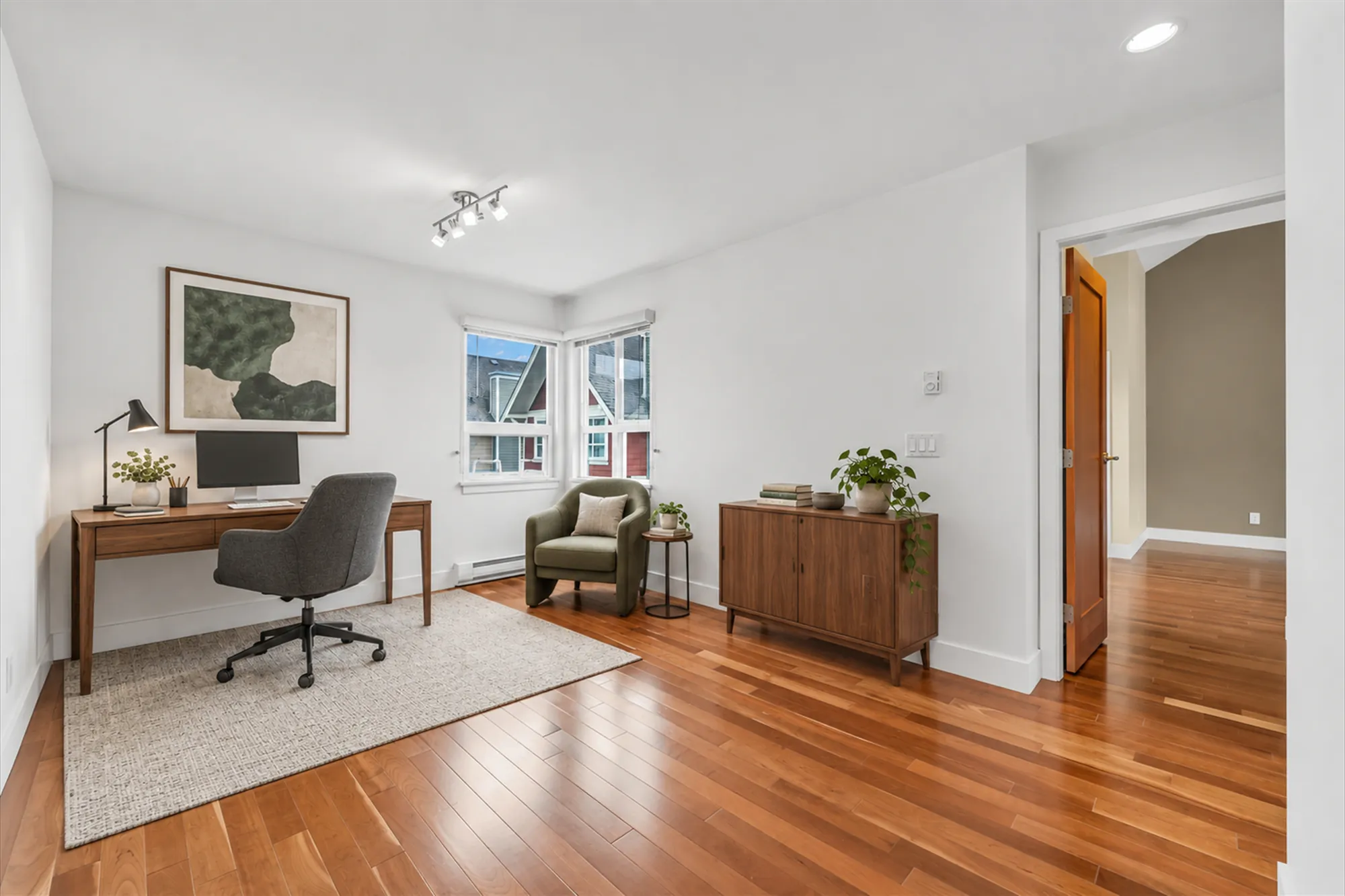 The same frame virtually staged as an office: a walnut desk with a lamp and a monitor, an office chair, a green armchair, a side table, a cabinet, a rug, a plant, an abstract print