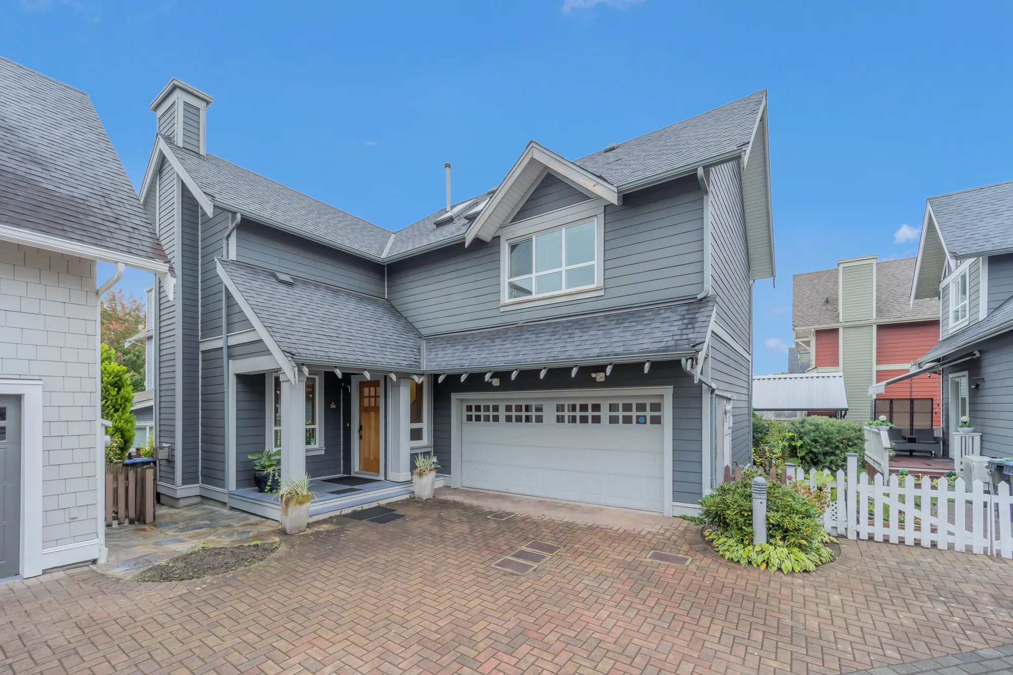 The front of 264 Holly Avenue from the brick-paved court: two storeys in grey siding with white trim, a covered porch with a wood front door, a white double garage door and a white picket fence