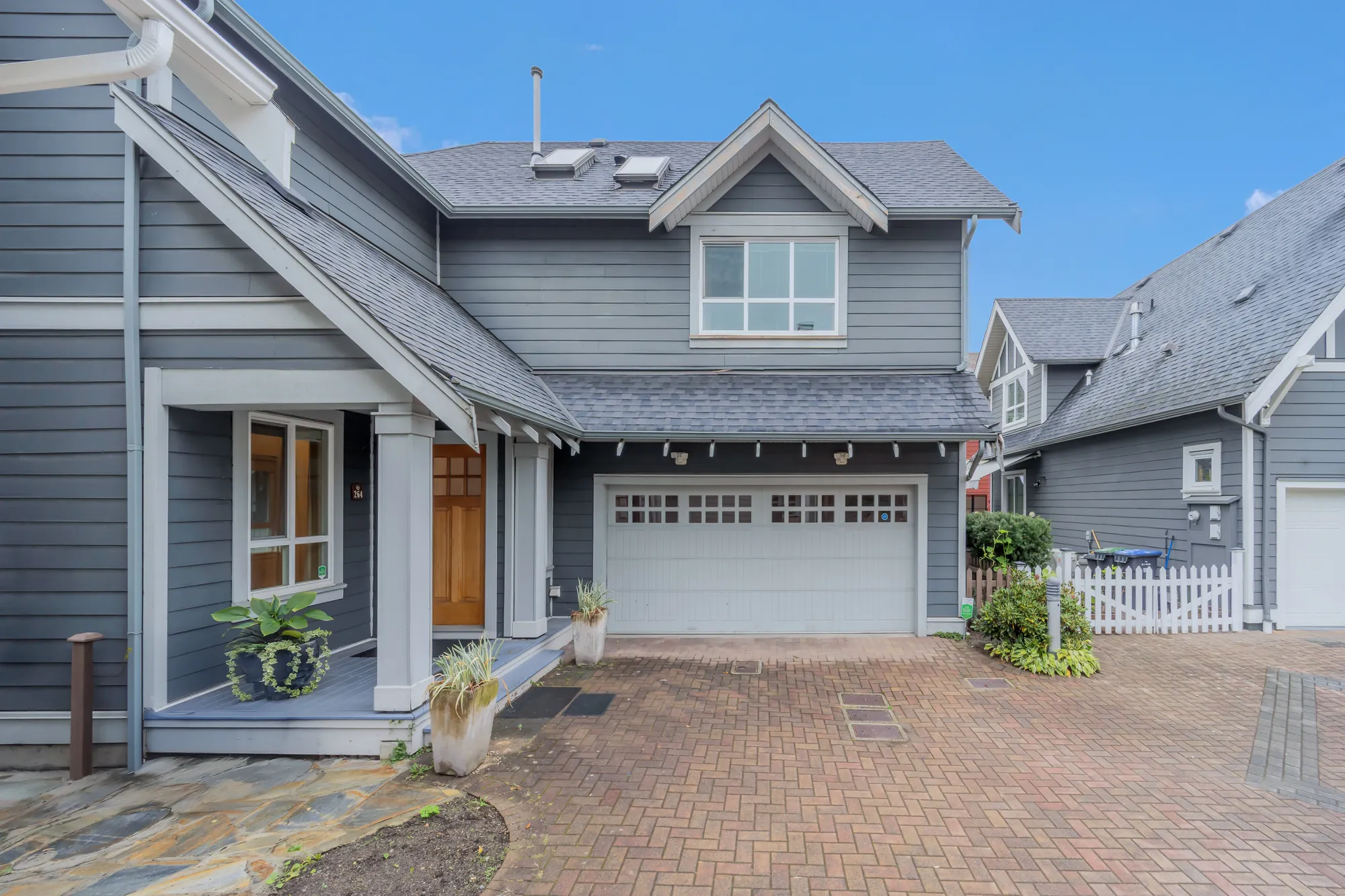The front from the other side: the covered porch on white columns, a large window and the wood front door, the white garage door, a lamp post and a white picket gate at the right