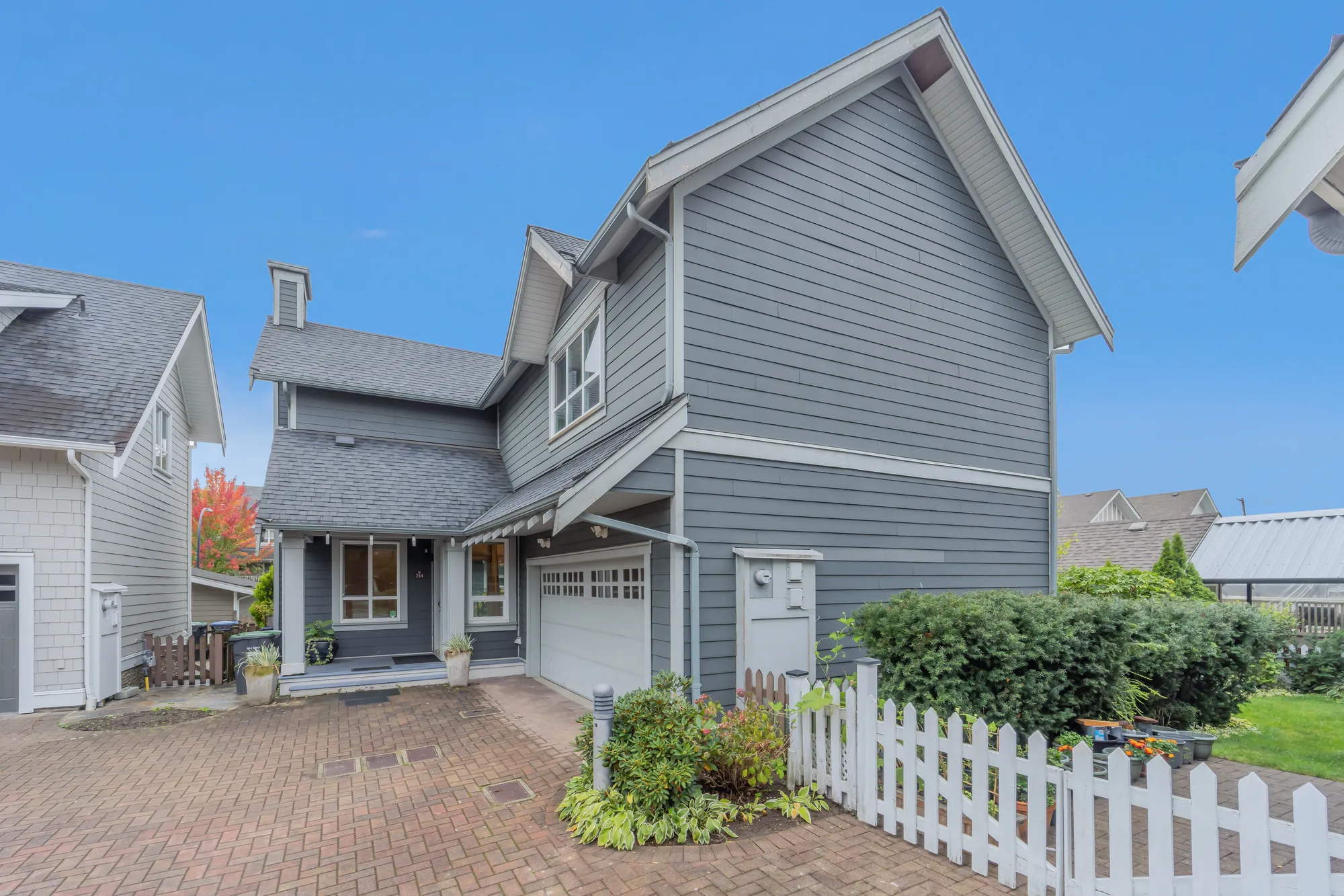 The house from the corner of the court: the porch and the garage door, the tall side wall under a gable, a white picket fence along a planted bed