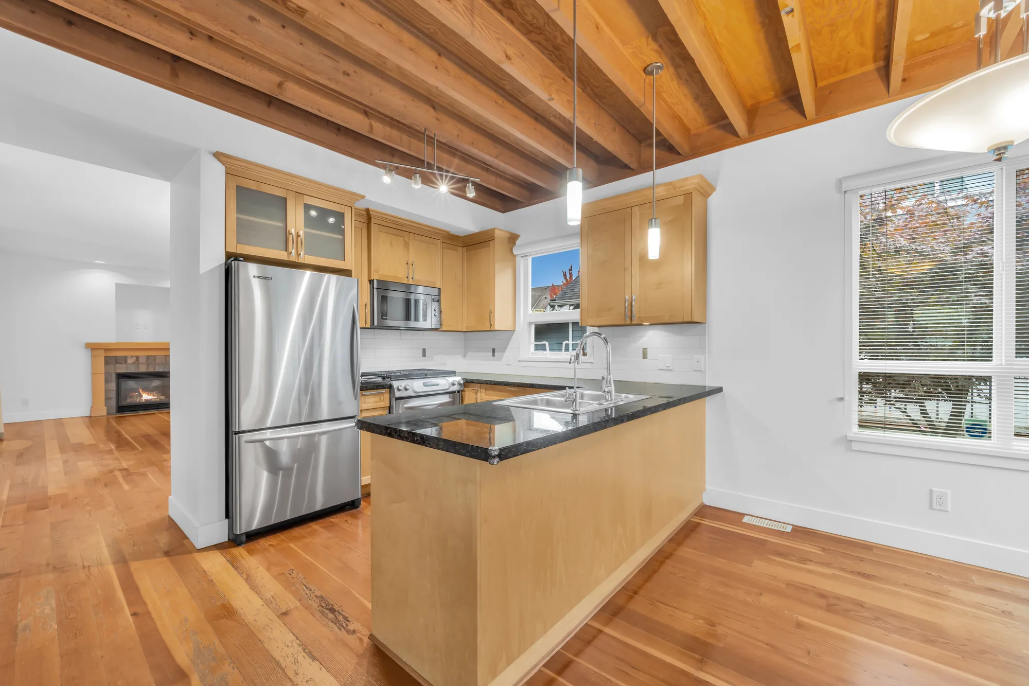 The kitchen: maple shaker cabinets with glass-front uppers, a stainless refrigerator and microwave, a range, a sink in a peninsula topped in dark granite, exposed wood joists overhead, the living room fireplace through the doorway at the left
