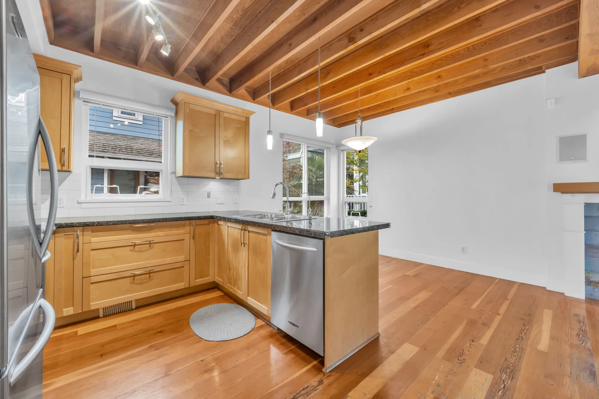 The kitchen from beside the refrigerator: maple cabinets on three sides, a dark granite counter, a dishwasher, a sink under a window, pendant and track lights under the wood joists