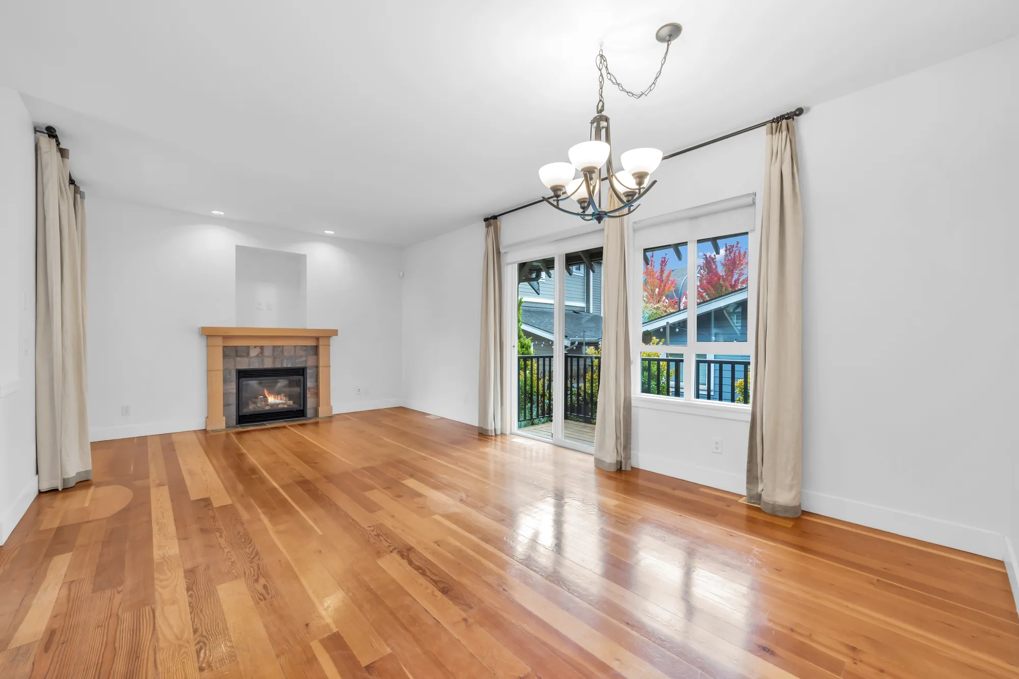 The living and dining room, empty: wood floors, a fireplace in slate tile with a wood mantel and a niche above it, sliding doors and a window with long curtains, a chandelier