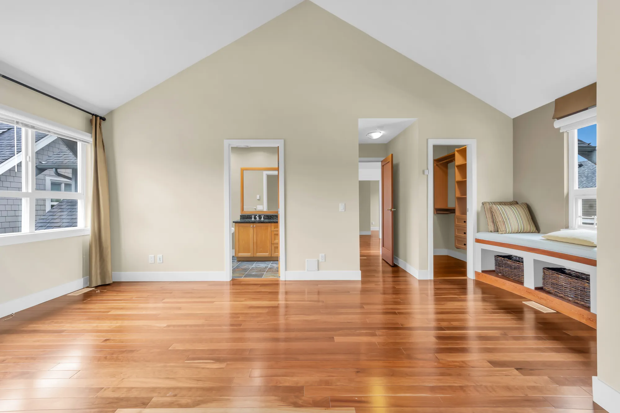 The primary bedroom under a vaulted ceiling: wood floors, a wide window with bronze curtains at the left, the doors to its bathroom and the hall, a walk-in closet, a window seat with cushions and baskets at the right