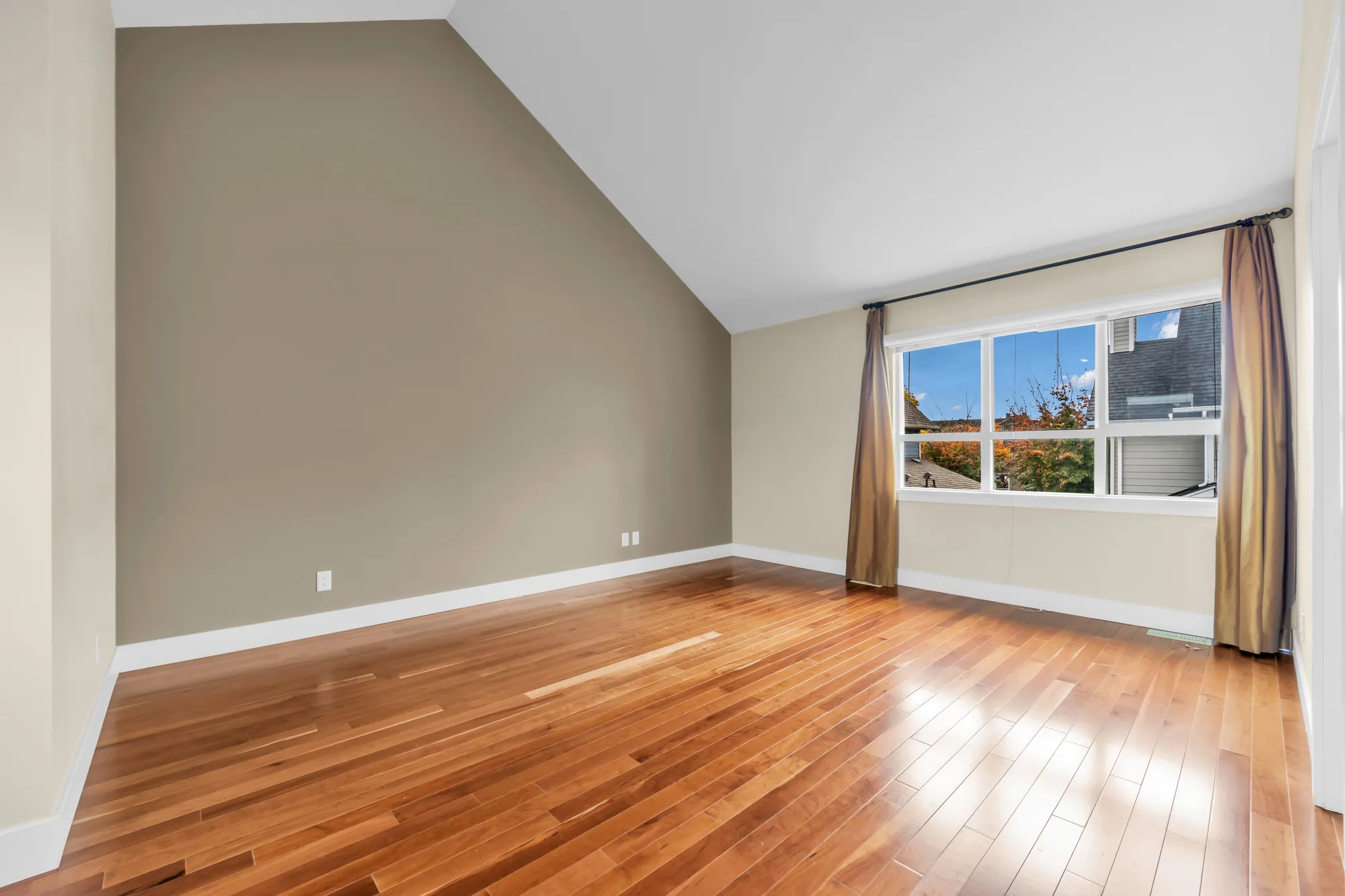 The primary bedroom, empty: a taupe wall under the vault, a wide window with bronze curtains, wood floors