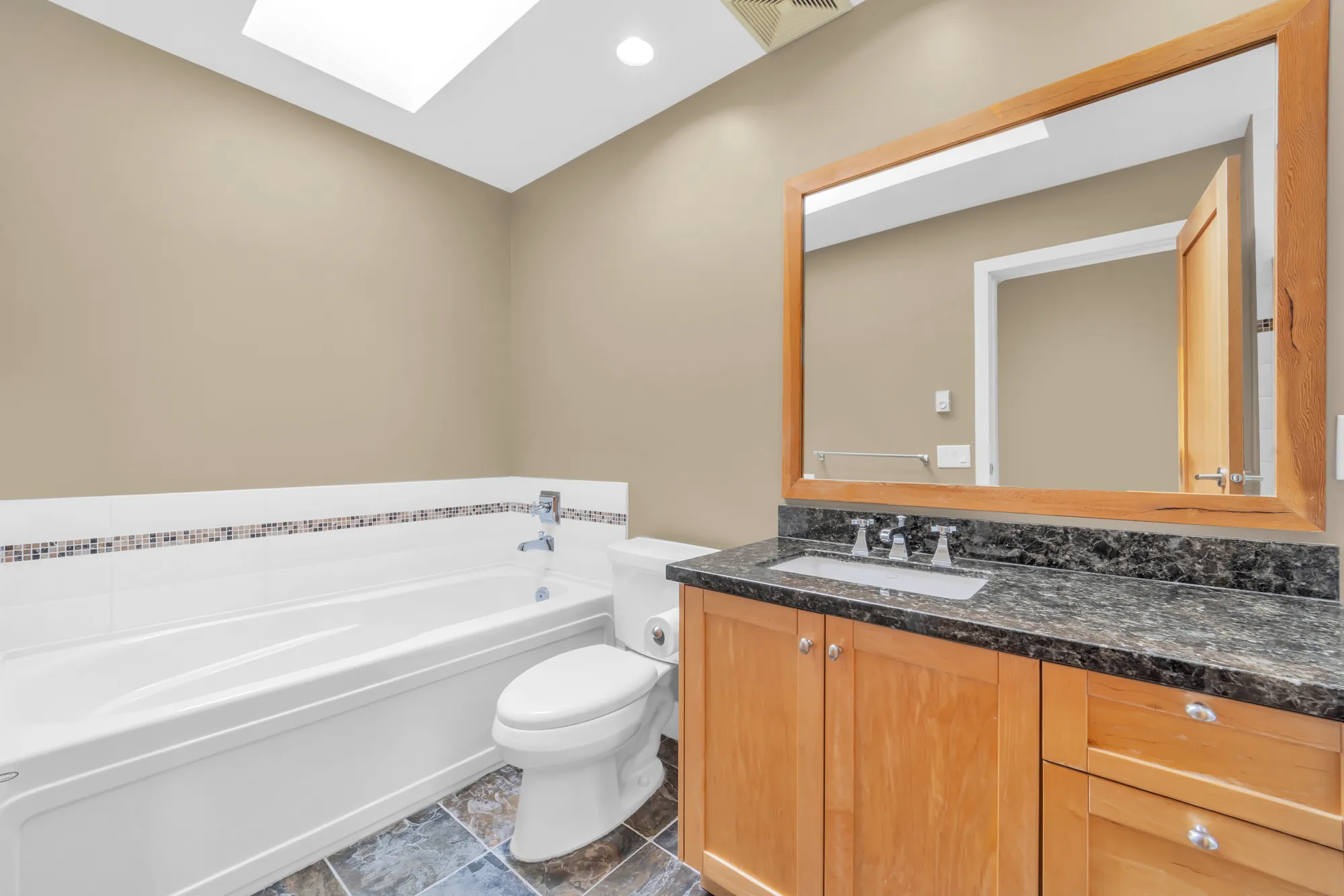 The primary bathroom: a soaker tub with a mosaic border, a toilet, a maple vanity with a dark granite top and a wood-framed mirror, a skylight
