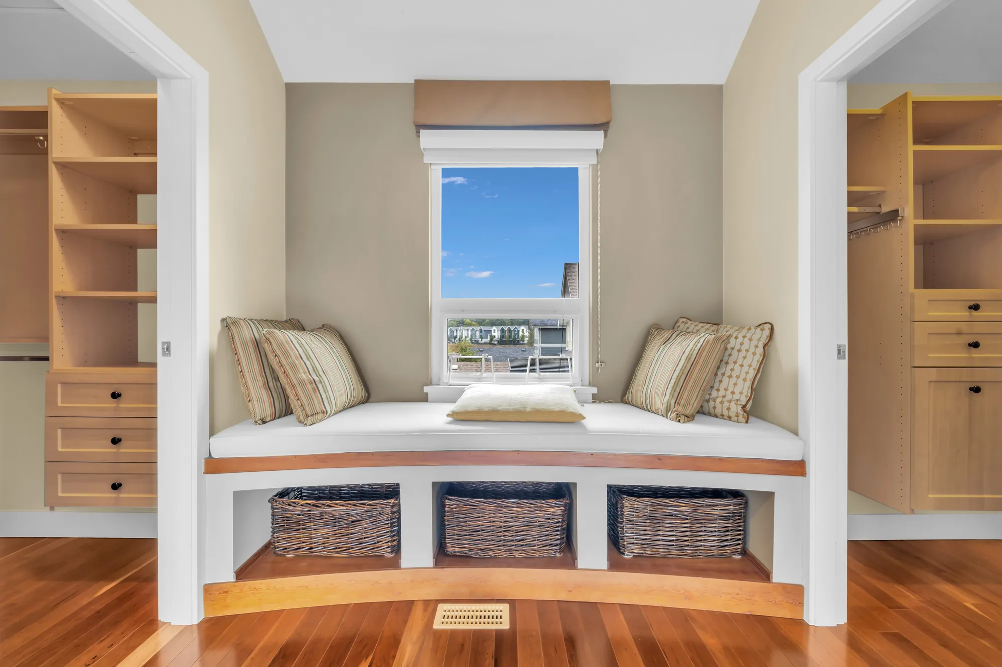 A window seat with a white cushion, pillows and three baskets below it, under a window, between the open doors of two walk-in closets fitted in maple