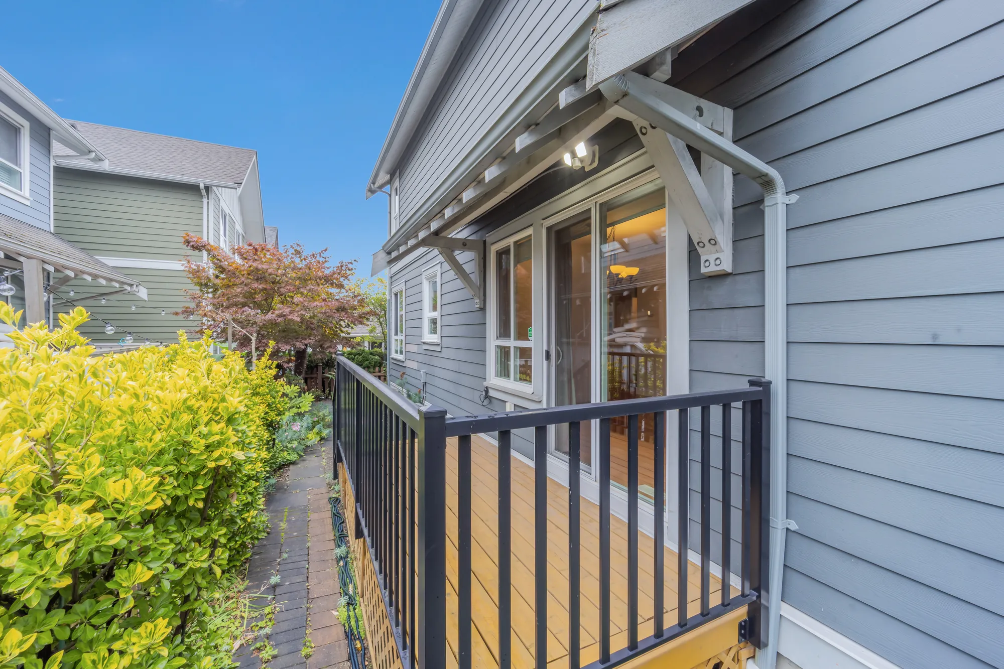 A narrow wood deck along the side of the house with a black railing, a sliding door with a chandelier lit inside, yellow-leaved shrubs at the left